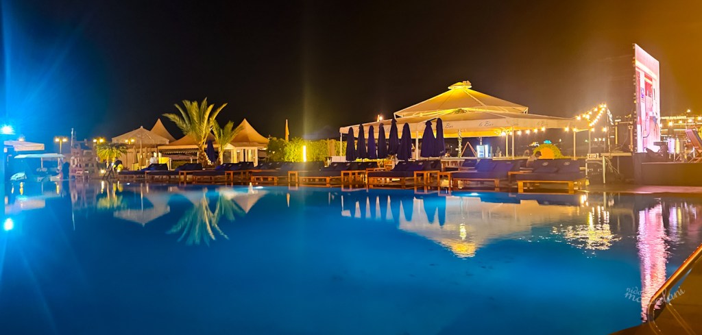 Night view of the pool area at The Saint Georges Hotel, featuring illuminated surroundings, lounge chairs, and palm trees reflecting in the water.